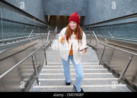 Photo en extérieur d'une jeune femme qui planifie un itinéraire, suit la carte sur l'application pour smartphone, monte les escaliers avec un sac à dos et sourit Banque D'Images