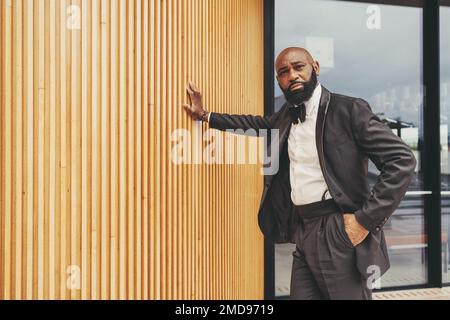 Un homme noir galant avec un drool et une tête chauve, portant un tuxedo, un noeud noir, et une chemise blanche; a une main dans sa poche et l'autre repose-main Banque D'Images