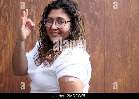 Une femme latine adulte avec un chemisier blanc cheveux ondulés et des lunettes montre son bras avec un bandage parce qu'elle vient d'être vaccinée contre Covid-19 Banque D'Images