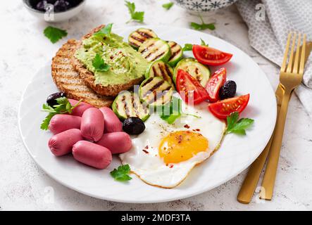 Œuf frit avec mini-saucisse, tomates, courgettes grillées, olives noires et toasts avec guacamole. Petit déjeuner anglais. Brunch. Banque D'Images