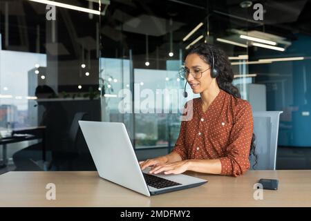 Femme d'affaires hispanique souriante travaillant au bureau avec un ordinateur portable et un casque pour les appels vidéo, femme assise sur le lieu de travail heureuse de travailler avec les clients. Banque D'Images