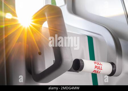 Poignée de porte de sortie de secours dans un avion volant avec rayons du soleil depuis une petite fenêtre, vue rapprochée. Banque D'Images