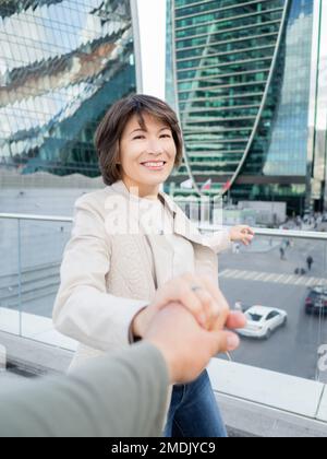 L'homme mène la femme souriante à la main contre le fond des murs en verre des bâtiments modernes. Centre-ville avec gratte-ciel et centres d'affaires financiers. Banque D'Images