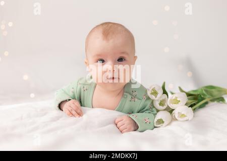 bébé mignon dans un body en coton vert à la maison sur un lit blanc avec des tulipes. Printemps, Fête des mères, 8 mars. Photo de haute qualité Banque D'Images