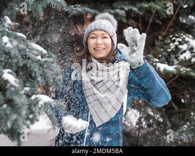 Femme souriante dans le chapeau tricoté de câble joue avec la neige. Amusez-vous dans le parc entre des sapins enneigés. Une femme rit alors qu'elle lance un ballon de neige. Saison froide. Banque D'Images