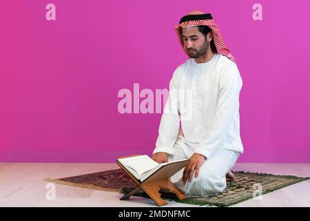 Jeune homme musulman arabe en vêtements traditionnels lisant le livre Saint Coran sur le tapis de prière avant un dîner iftar lors d'un festin de ramadan à la maison Banque D'Images