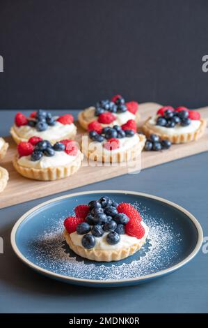 Plusieurs petites tartes de crème anglaise à la vanille sur une planche à découper en bois derrière une tarte placée sur une assiette bleue. Banque D'Images