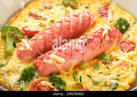 Omelette ou frittata avec deux saucisses, fromage fondu, tomates cerises, brocoli et persil sur poêle rouge ou poêle en fer sur fond blanc en béton Banque D'Images