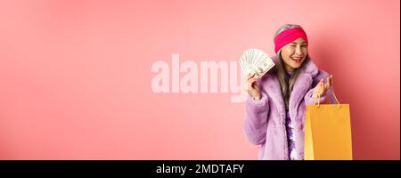 Femme asiatique riche et à la mode gaspillant de l'argent dans les magasins, tenant sac de shopping et dollars, en se faisant un plaisir à l'appareil photo, fond rose Banque D'Images