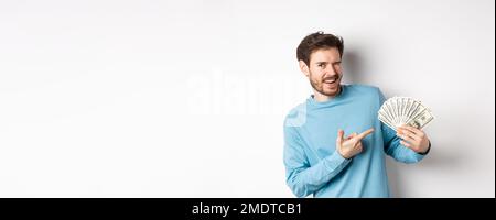 Beau gars riche qui se vante à la caméra, montrant des revenus, pointant le doigt sur l'argent gagné et souriant, debout sur fond blanc Banque D'Images
