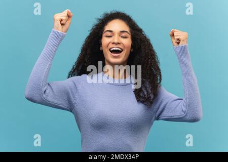 Portrait de la femme emballée excitée et habillée avec des mains levées et criant ouais, je suis gagnant, réjouissant victoire, succès. Prise de vue studio isolée sur fond bleu Banque D'Images