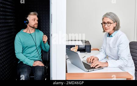 Examen audiologique. Audiologiste effectuant un contrôle auditif pour un homme adulte charmant dans un kiosque audiométrique insonorisé. Traitement de la perte auditive, vue latérale Banque D'Images