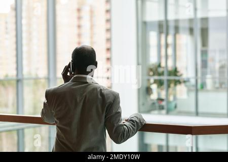 Vue arrière d'un directeur général afro-américain expérimenté parlant sur un téléphone portable tout en étant debout près d'une rampe dans le centre de bureau Banque D'Images