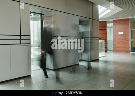 Partie d'un couloir spacieux à l'intérieur d'un centre de bureau moderne et deux hommes d'affaires flous par des ascenseurs avec des portes vitrées qui se déplacent le long Banque D'Images