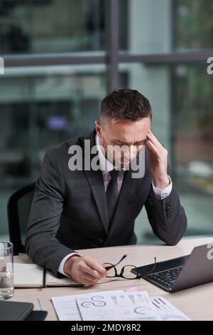 Le directeur général a surtravaillé avec la main sur le temple se sentant malade en étant assis devant un ordinateur portable sur le lieu de travail et en analysant les données en ligne Banque D'Images