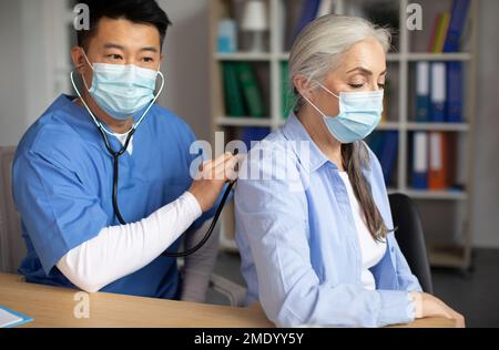 Grave homme coréen adulte médecin en masque de protection écoute avec stéthoscope à la respiration de la vieille dame européenne Banque D'Images