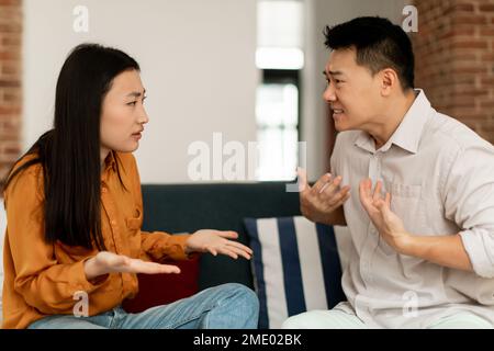 Les conjoints asiatiques en colère ont querelles, assis sur un canapé et argumentant, regardant les uns les autres, vue latérale Banque D'Images