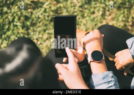 Heureux amis partageant du contenu multimédia en ligne avec leurs smartphones et SmartWatch, deux belles femmes Latina assis à l'extérieur Banque D'Images