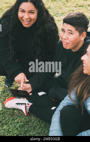 Groupe de Latinos riant assis sur le sol dans un parc avec un smartphone Banque D'Images