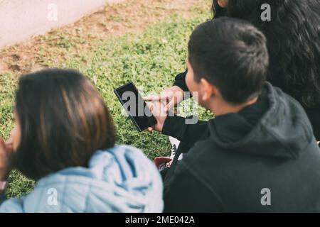 Vue depuis le groupe arrière de Latinos riant assis sur le sol dans un parc avec un smartphone Banque D'Images