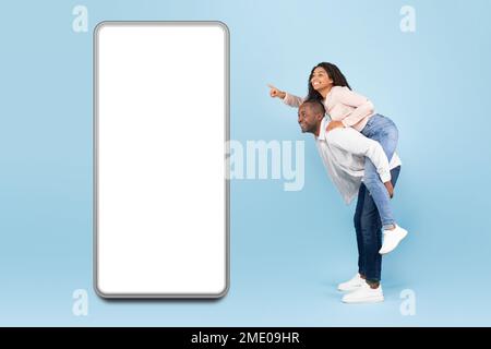 Application mobile exceptionnelle. Couple noir présentant un grand smartphone géant avec maquette, homme de la femme sur son dos Banque D'Images