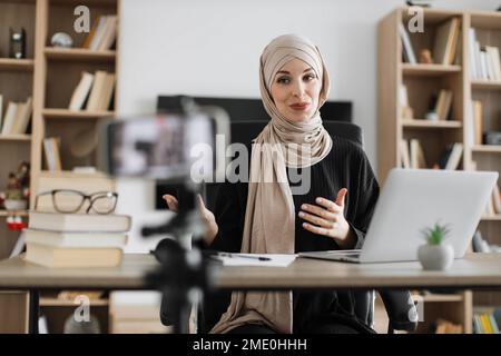 Charmante femme musulmane dans hijab parlant et gestant tout en enregistrant la vidéo sur le téléphone moderne. Femme blogueuse assise à la maison et faisant le flux en direct. Banque D'Images