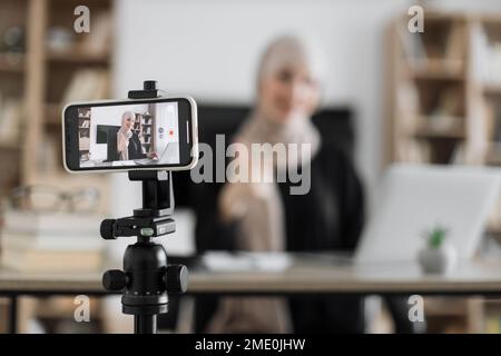 Concentrez-vous sur l'écran du téléphone de la femme arabe heureuse assise au bureau avec des livres, des notes et un ordinateur portable parlant avec des suiveurs. Une femme qui travaille à la maison pendant l'apprentissage à distance et montre le pouce vers le haut. Banque D'Images