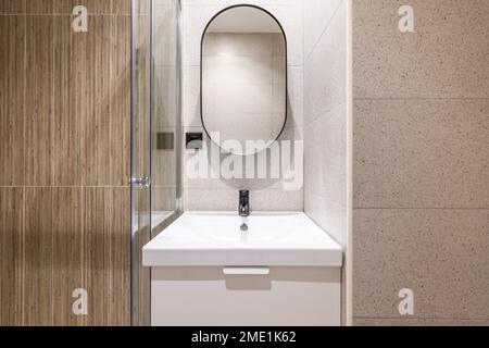 Salle de bains avec éclairage artificiel lumineux, meuble-lavabo en céramique blanche et solide. Grand miroir ovale moderne sur le mur au-dessus de l'évier. Banque D'Images