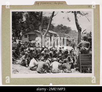 Photographie d'un groupe de prisonniers japonais gardés par la police militaire américaine. JAP prisonniers Huddle pour Chow on Saipan. Capturé dans le balayage américain vers le nord au-dessus de l'île de Saipan, les prisonniers de Jap s'écraseront sur le sol et dévorent chamairement les rations passées de leurs magasins de capteurs. Les députés surveillent de près les soldats ennemis, en attendant leur retrait de l'île à bord d'un transport d'assaut assuré par la Garde côtière. Banque D'Images