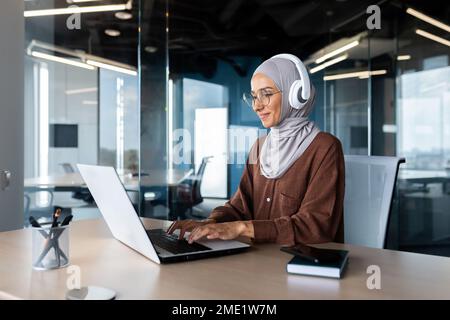 Formation en ligne. Jeune étudiante en hijab et casque, assise au bureau à la table, étudiant à distance avec un ordinateur portable. Saisie sur le clavier.sourire. Banque D'Images