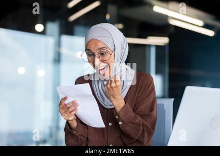 Jeune femme heureuse étudiante dans hijab. Assis à la table, tient une lettre, a reçu un avis d'admission à l'université, les résultats de l'examen. Célèbre, crie, montre le succès avec sa main. Banque D'Images