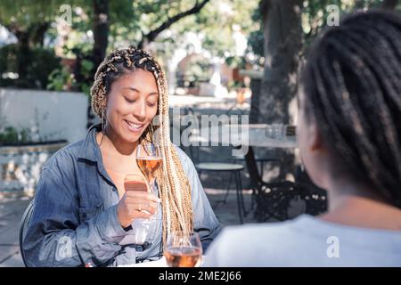 Souriant, de jeunes amies ethniques avec des tresses assises à table et buvant du vin mousseux le jour ensoleillé dans le parc tout en regardant les uns les autres Banque D'Images