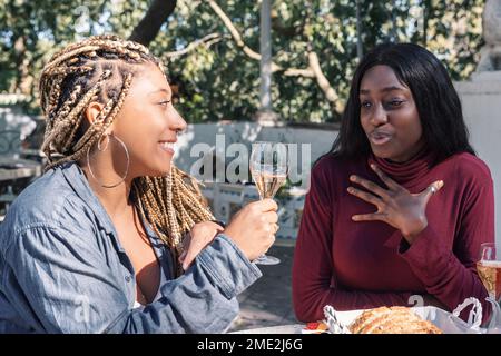 Souriant, de jeunes amies ethniques avec des tresses assises à table et buvant du vin mousseux le jour ensoleillé dans le parc tout en regardant les uns les autres Banque D'Images