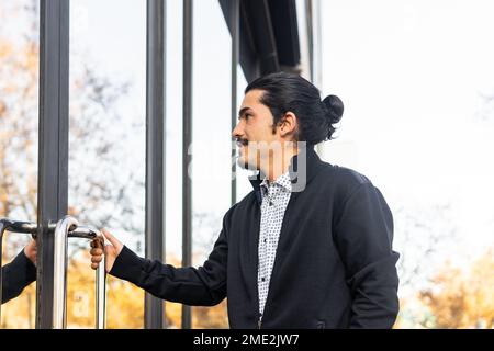 Vue latérale d'un jeune homme hispanique à barbe confiant, vêché de vêtements élégants et décontractés, ouvrant la porte d'un bâtiment moderne avec des murs en verre et miroirs ensoleillés Banque D'Images
