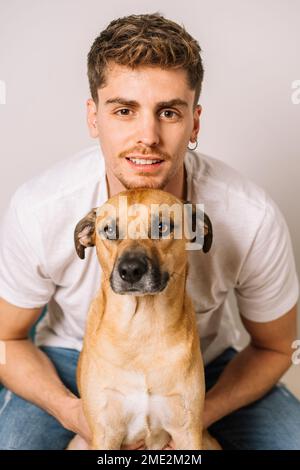 Un homme souriant avec une chemise blanche et un Jean assis derrière un adorable chien de race et un petit câlin tout en regardant l'appareil photo Banque D'Images
