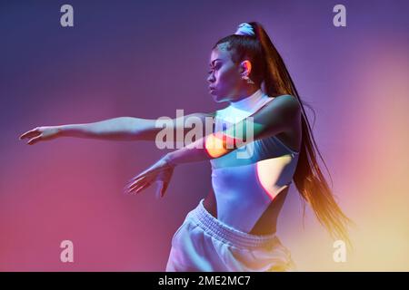 Jeune danseuse talentueuse avec longue queue de cheval droite en body blanc qui se déplace le corps et s'étend les bras sur fond coloré Banque D'Images
