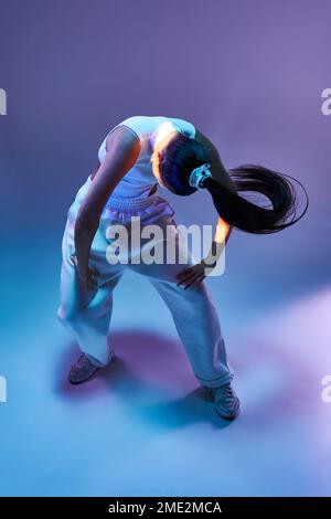 Le corps de la jeune danseuse en pantalon sportif se penche vers l'avant tout en musique contre les néons violets Banque D'Images