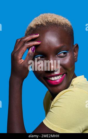 Positive Afro-américaine femme avec cheveux courts et maquillage touchant le visage et regardant loin tout en souriant sur fond bleu Banque D'Images