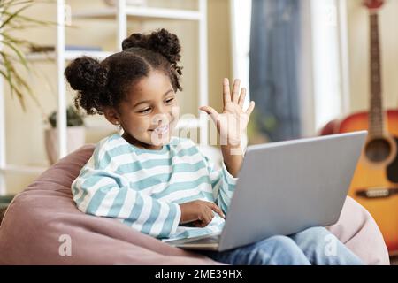 Portrait d'une petite fille noire agitant à l'appareil photo d'ordinateur portable et souriant tout en appréciant la leçon en ligne de la maison assis dans confortable chaise de sac de haricot, espace de copie Banque D'Images