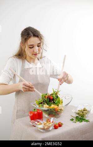 jeune fille adolescente agite diligemment salade penchée sur un bol souriant elle aime cuisiner des aliments sains ingrédients végétariens feuilles de laitue verte cuillères en bois tablier et nappe beige couleur Banque D'Images