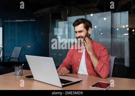 Un employé de bureau a un mal de dents, un homme d'affaires malade travaille à l'intérieur du bureau avec un ordinateur portable, un homme a un mal de dents sur le lieu de travail. Banque D'Images