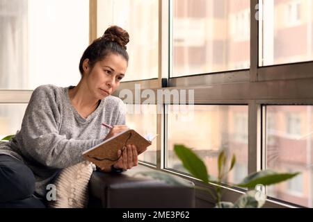 Femme écrivant un journal intime dans un canapé à l'intérieur entouré de plantes Banque D'Images