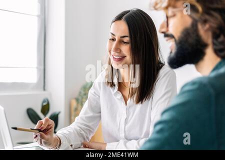 Femme d'affaires souriante pointant vers un ordinateur portable devant un collègue Banque D'Images