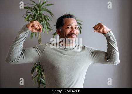 Homme mûr heureux fléchissant les muscles devant le mur gris Banque D'Images