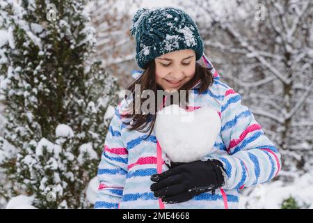 Fille souriante tenant un boule de neige en forme de cœur en hiver Banque D'Images