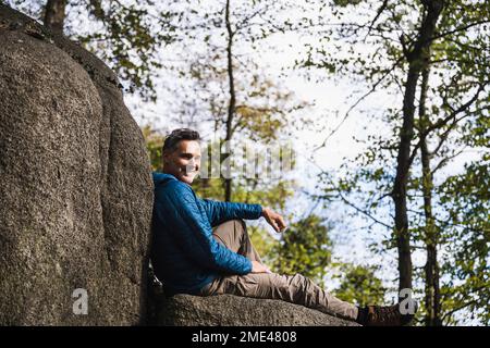 Homme mûr souriant assis par le rocher devant les arbres Banque D'Images