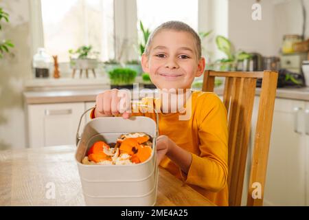 Garçon souriant montrant des peaux de mandarine dans un seau à compost à la table à manger Banque D'Images