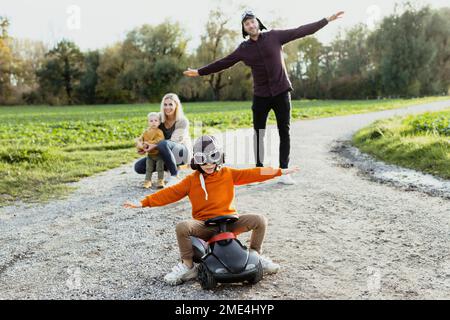 Garçon portant des lunettes volantes assis avec les bras tendus sur la voiture bobby devant la famille Banque D'Images