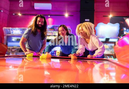 Amis multiraciaux s'amusant à jouer au hockey pneumatique dans une salle de jeux d'arcade Banque D'Images