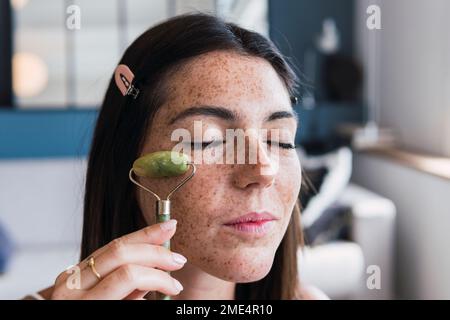 Jeune femme avec le visage freckle massant à l'aide du rouleau de pierre de jade à la maison Banque D'Images